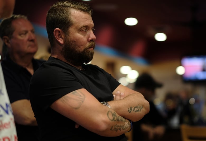 Two men with serious expressions on their faces and their arms crossed stand and listen to a speech.