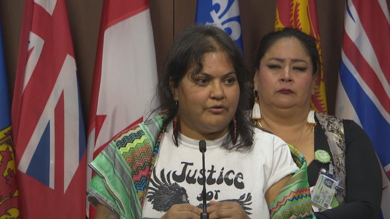 A South Asian woman speaks in front of flags, as an Indigenous woman looks on.