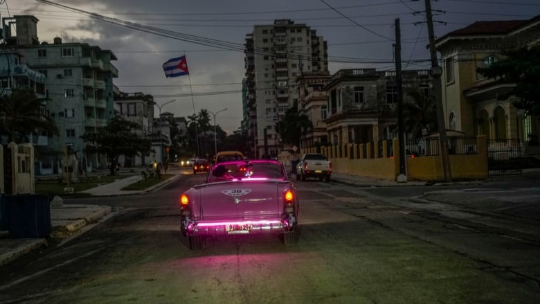 A classic American car with a Cuban flag attached to it, drives along a Havana street.