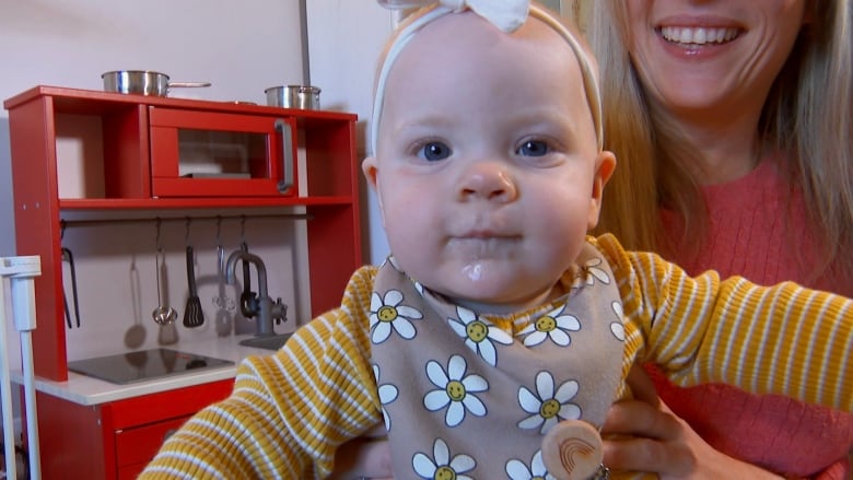 A baby wears a striped yellow and white onesie and a bib with daisies on it. She also wears a white bow on her head. 