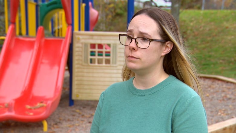 A woman wears glasses and a turquoise shirt. She stands in front of a playground with a red slide. 