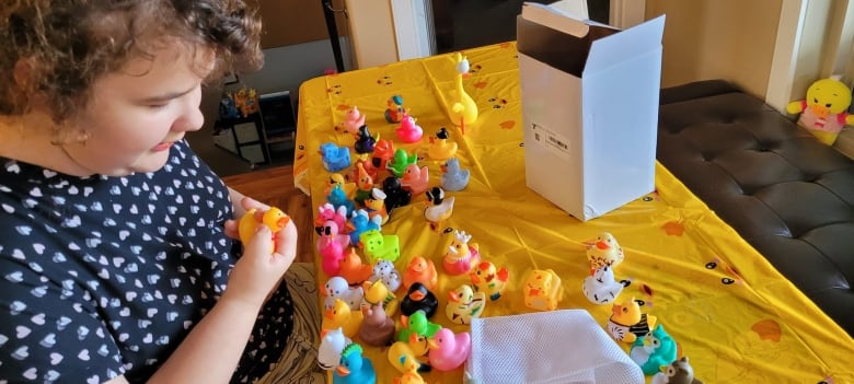 A dark-haired curly girl in a dark blue dress sits at a table covered in a yellow cloth, and filled with yellow ducks.