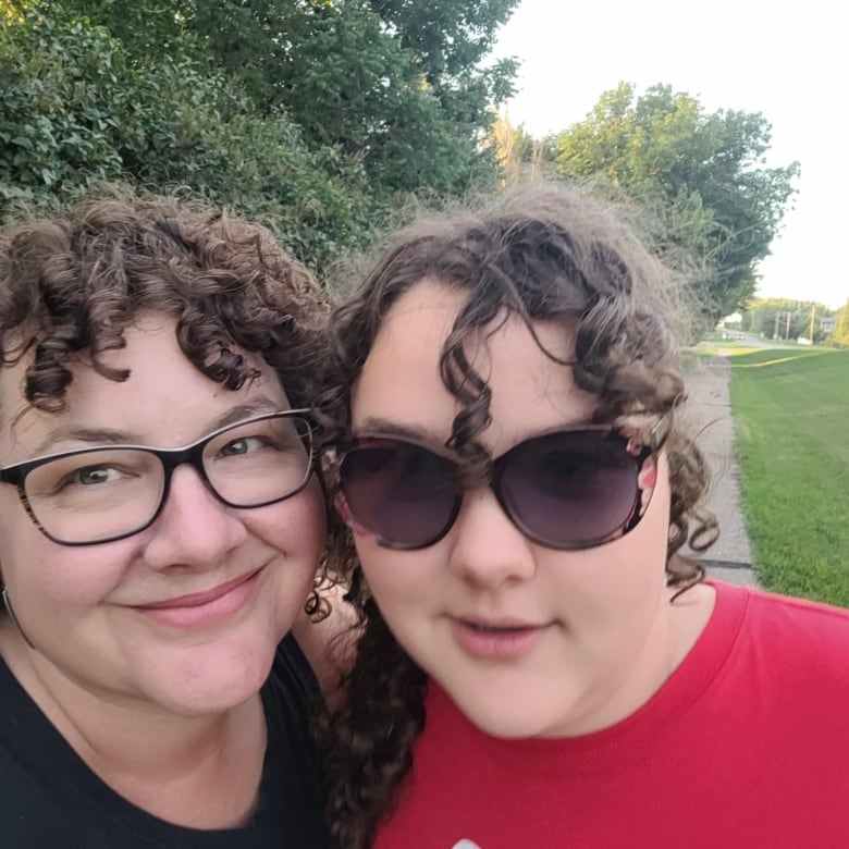 A mother and daughter with dark curly hairs stand with their heads close together, a park in the background. 