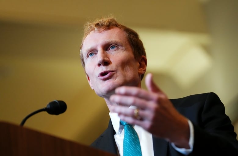 A man in a black suit and turquoise tie gestures with his hand as he speaks into a microphone.