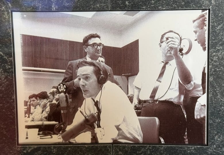 A black and white poto of a young man sitting behind a microphone that says CBZ. Other men are standing behind him.