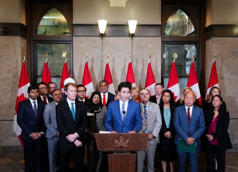 A man in a blue suit speaks at a podium as a number other people in business attire watch.