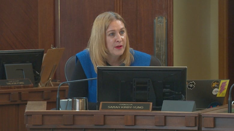 A woman with blond hair sits behind a council desk and speaks into a microphone.