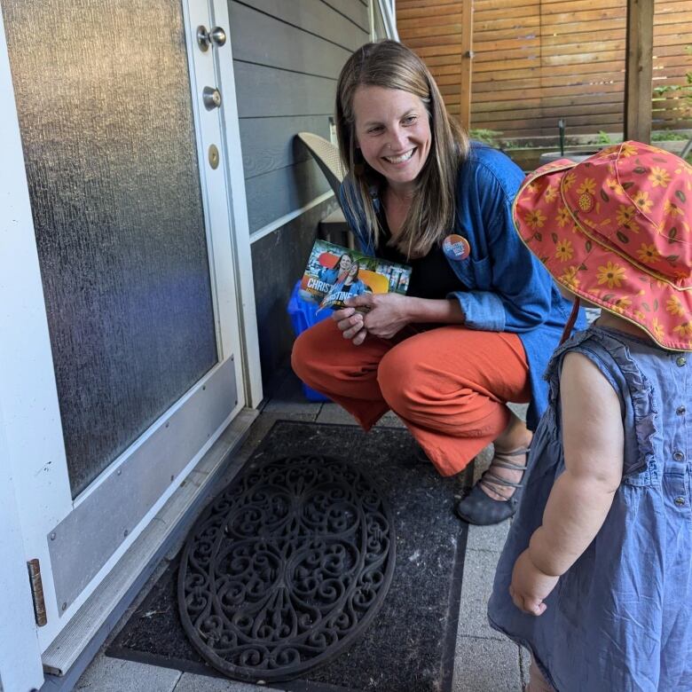 A woman smiles and crouches to speak with a toddler near the door of a residence.