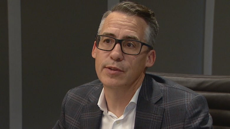 A man with greying hair wearing glasses a white button up shirt and a grey jacket