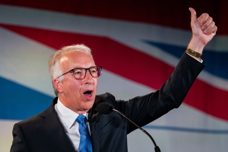 A man wearing a suit, his arm raised high in the air, gives a thumbs up.