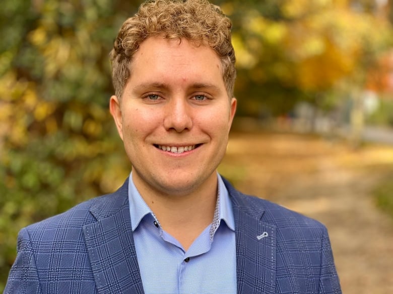 Man with blonde hair wearing a blue suite smiles for a photo in a park with bright autumn colours behind him.