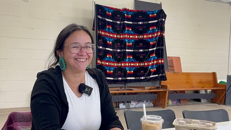 A brown-haired woman wearing a white shirt and black sweater and blue earrings sits at a table smiling.