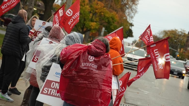 A group of people wearing red rain coats with the Unifor logo stand on the sidewalk in drizzling rain on a grey morning