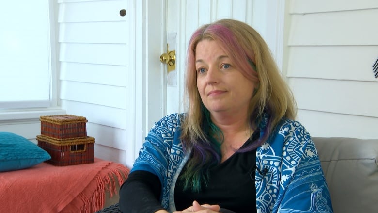 A woman sits on a couch. She wears a blue and white patterned cardigan. She has blonde hair with pink and green streaks. 