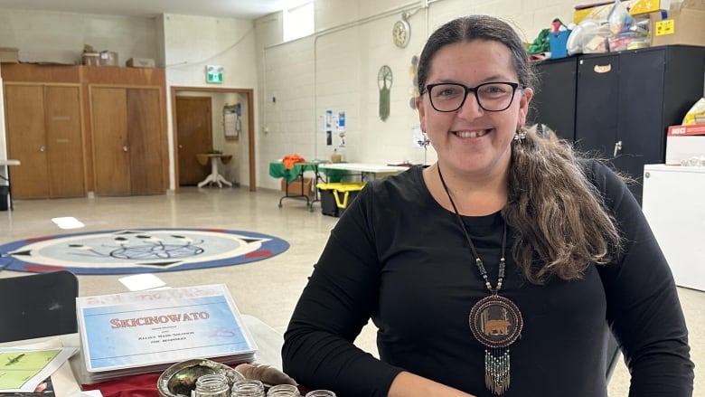 A bespectacled woman with long brown hair pulled into a pony tail and wearing a black shirt and beaded necklace sits at a table smiling.