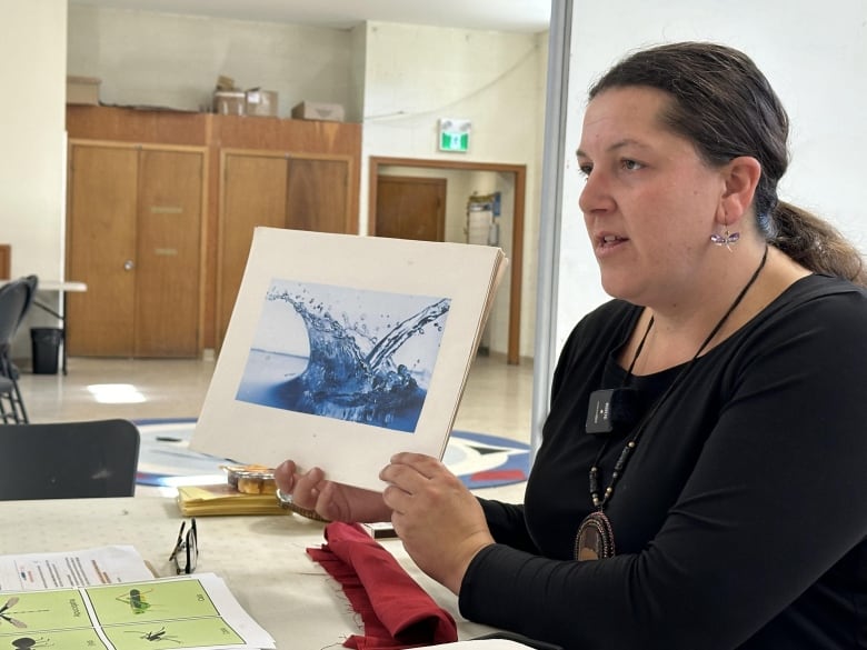 A woman with long brown hair pulled into a pony tail sits at a table and holds up a photo of splashing water.