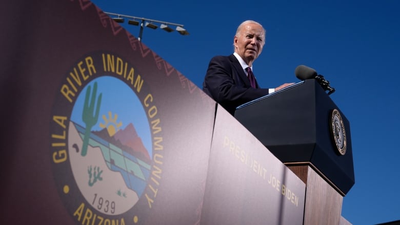 A man in a suit and dark aviator sunglasses stands at a podium on a sunny day.