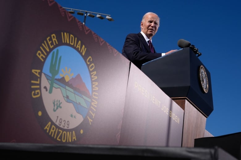 A man in a suit and dark aviator sunglasses stands at a podium on a sunny day.