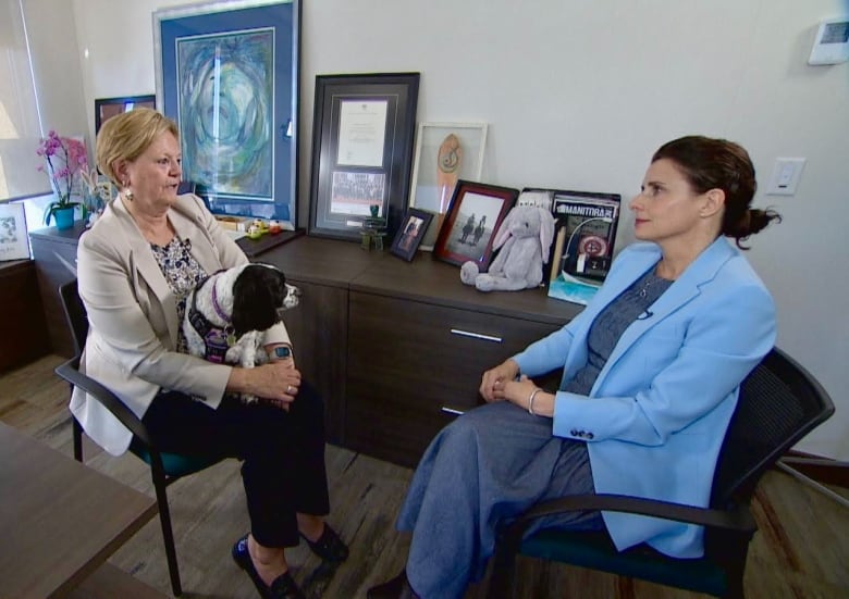 A woman with short blond hair wearing a beige blazer, black trousers, and black loafer style shoes sits in an office chair with a small black and white dog in her lap. The dog is wearing a black harness. Across from her sits a woman with black hair in a ponytail wearing a dark blue dress and light blue blazer. The women are facing each other.