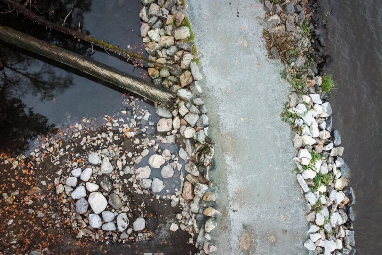 A drone shot of a bouldered pathway with a log, a small pond on the left, and the slightly visible water of a much larger lake on the right. 