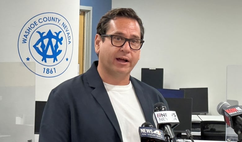 A man with dark hair and glasses in a navy blazer and white t-shirt speaks into microphones in an office setting. 