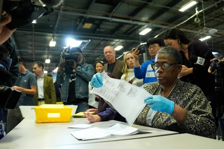 A person with glasses wearing blue latex gloves sits at a table holding a paper ballot as members of the media look on. 