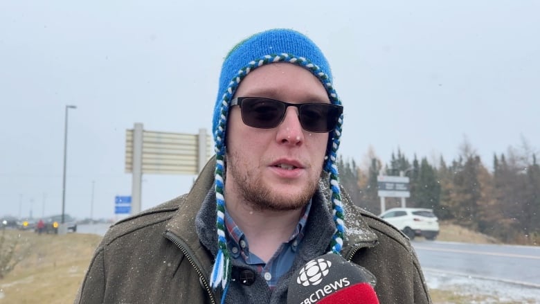 Man in sun glasses wearing a blue hat that comes down over his ears. He's wearing a brown jacket and it's lightly snowing.