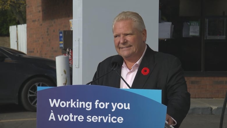 a man in a dark blazer and a white shirt with an open collar, wearing a poppy, stands at a podium that says 