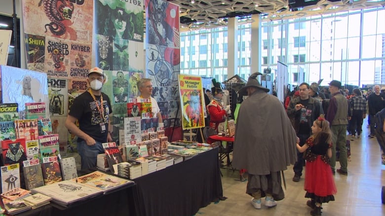 A father and daughter dressed up in costumes look at a booth of comics. 