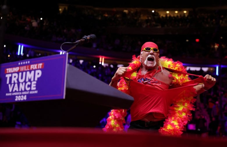 A performer wearing a bandana, sunglasses and a boat scarf rips off their shirt while speaking onstage.