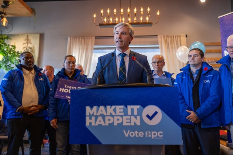 Man in blue suit stands at blue podium with several people standing at his side