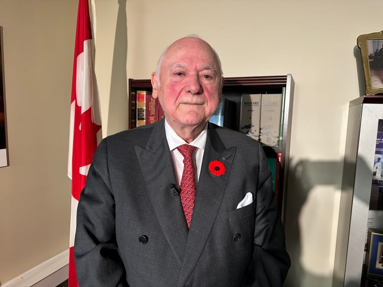 A man in a suit with a poppy stands in front of a Canadian flag