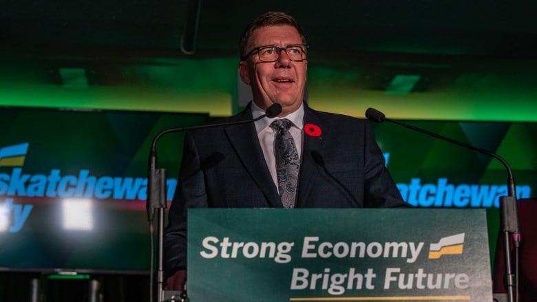 A man wearing glasses and a poppy in his lapel, stands at a podium, bearing the words: Strong economy, Bright future.