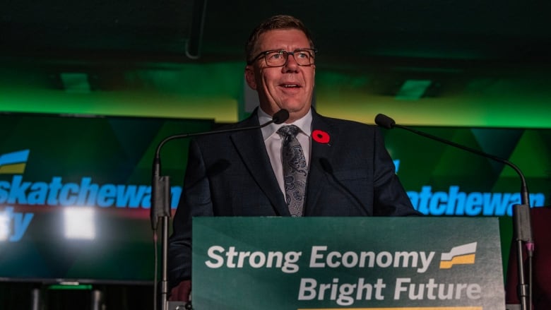 A man wearing glasses and a poppy in his lapel, stands at a podium, bearing the words: Strong economy, Bright future.