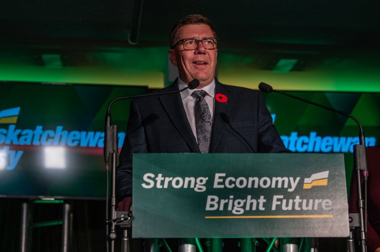 A man wearing glasses and a poppy in his lapel, stands at a podium, bearing the words: Strong economy, Bright future.