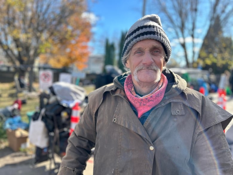 A man with a hat, scarf and brown jacket grins at the camera with fall leaves and a shopping cart in the background. 