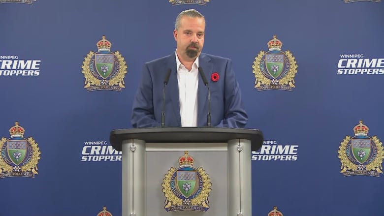 A man wearing a blue suit and a red poppy stands at a podium to speak. 