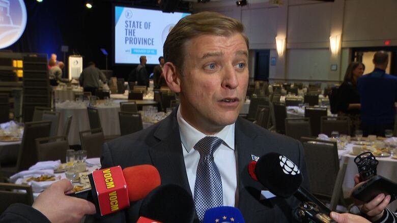 A man wearing a suit stands among set tables at a business conference. He's speaking into several microphones held by reporters.