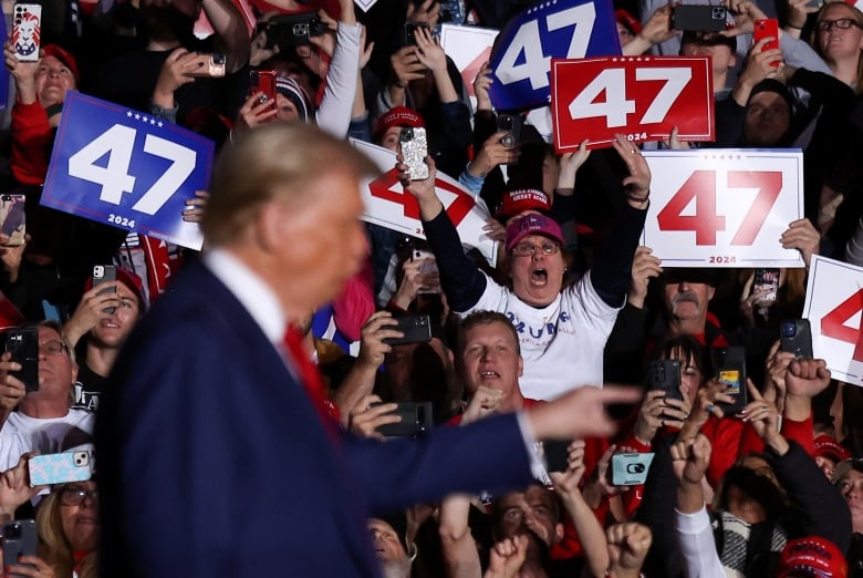 An excited woman raises both her arms in the air in a crowd during a political rally. A man in a suit is out of focus on stage.