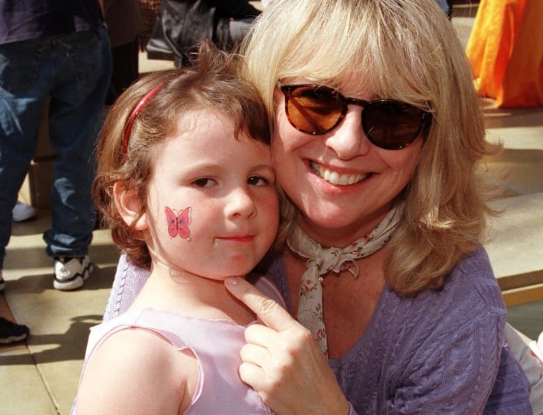 A woman wearing sunglasses and a mauve sweater kneels next to a young girl. 