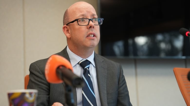 A man in a suit sits a table with a microphone.