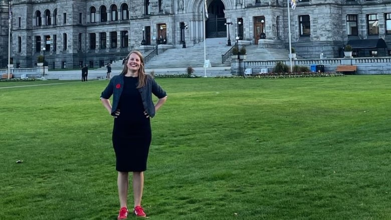 A woman in a black dress with red shoes stands on green grass with the B.C. legislature in the background.