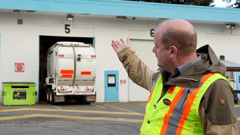 A man in a safety vest gestures at a parking bay that can barely fit the garbage truck parked there with its end hanging out.