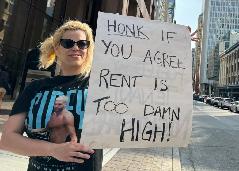 A blonde woman holds a sign that says 'Honk if you agree rent is too damn high!'