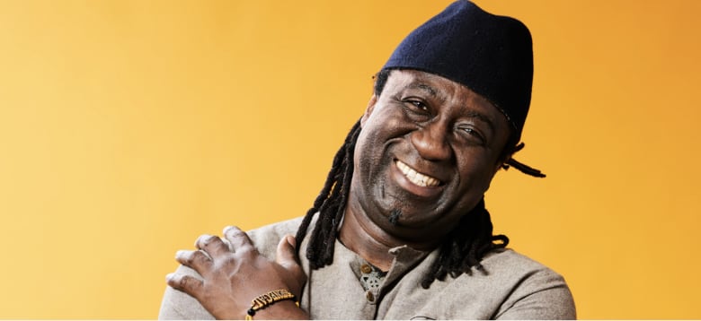 A man with a broad smile, black beard and shoulder-length dreadlocks falling from his black hat. He has one hand on his right shoulder. His photo against a yellow background.