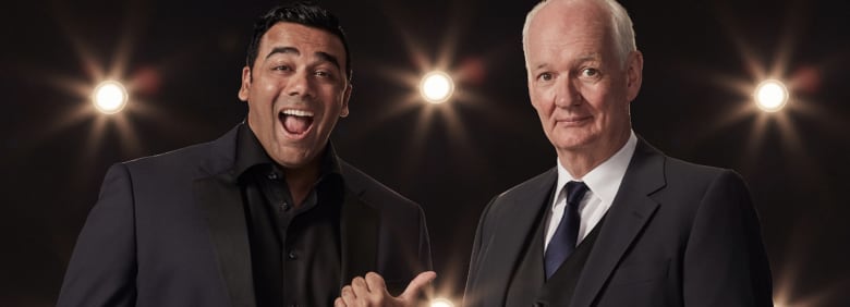 Two men, one smiling with his mouth open with black hair and the other with white hair on the sides on stagw with lights in the background. Both in black suits.