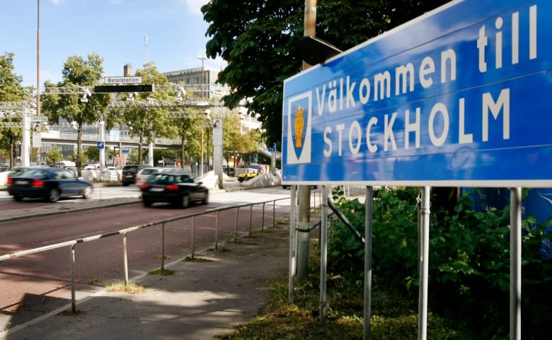 Cars on a highway on the left pass under an arch with cameras on the left. On the right a big blue sign reads 'Valkommen till Stockholm.'