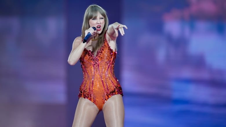 A woman stands on a stand in a red bodysuit singing and pointing at the crowd. 