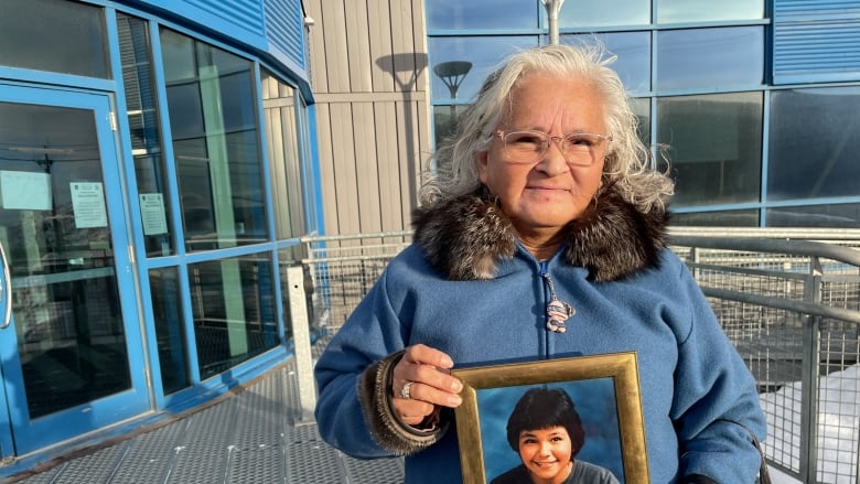 A woman wearing a parka with white hair holds a framed photograph of a young girl