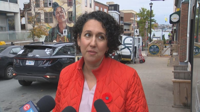 A woman with short, dark curly hair standing in front of mics while on a sidewalk. She is wearing a red jacket with a poppy pin.